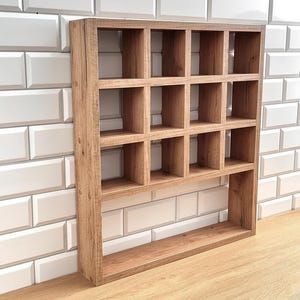 May include: A wooden wall-mounted display shelf featuring a light brown wood grain finish. The unit consists of nine square compartments arranged in a three-by-three grid at the top, with a single wider rectangular shelf space located at the bottom.