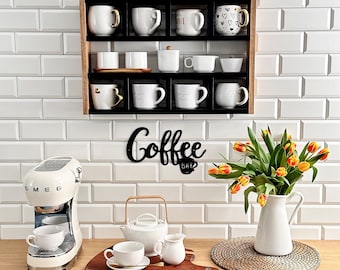 Può includere: Una mensola da parete in legno nero a tre ripiani contenente varie tazze in ceramica bianca, una macchina da caffè Smeg color crema, una teiera bianca, una lattiera, un tagliere in legno e una brocca bianca riempita di tulipani arancioni e gialli.