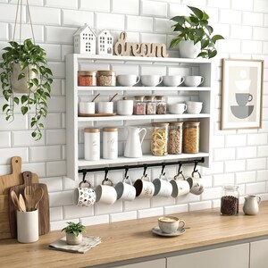 Peut inclure: Une étagère de cuisine murale blanche à trois niveaux contenant divers bocaux en verre, des tasses en céramique blanche et un pichet blanc. Sous l'étagère, une barre en métal noir supporte six tasses suspendues. Une enseigne en bois indique dream.