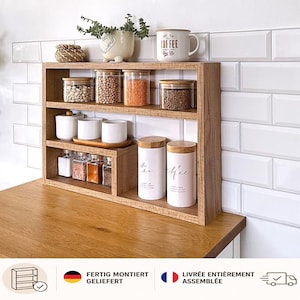 Può includere: Uno scaffale da banco in legno a tre livelli contenente vari barattoli di vetro con coperchi in bambù, contenitori in ceramica bianca con le scritte tea e coffee e una piccola pianta in vaso, organizzati con prodotti secchi e spezie.