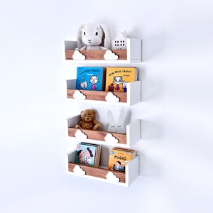 Pode incluir: Quatro prateleiras flutuantes brancas e de madeira com recortes de nuvens. Cada prateleira contém livros infantis e bichos de pelúcia, incluindo um coelhinho branco, um ursinho de pelúcia e uma casa branca. Os livros têm capas coloridas.