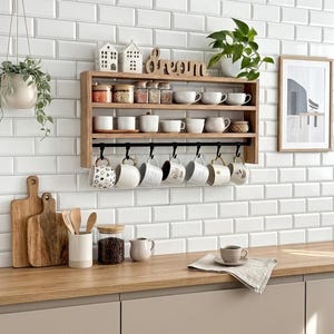 Może przedstawiać: Dwupoziomowa drewniana półka ścienna z czarną metalową szyną i siedmioma haczykami, na których wiszą ceramiczne kubki. Na półce znajdują się szklane słoiki, białe ozdoby w kształcie domków, drewniany napis "dream" oraz zielona roślina.