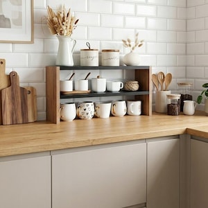 May include: A two-tier countertop shelf with black metal shelves and light wood side supports. The shelf holds white ceramic mugs, some with gold accents or heart patterns, and small white canisters labeled sugar and salt, arranged on a wooden kitchen counter.