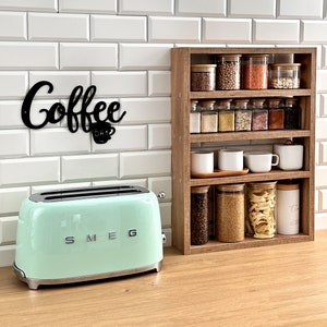Peut inclure: Un grille-pain vert clair avec le mot "SMEG" sur le devant, posé sur un comptoir à côté d'une étagère en bois avec des bocaux en verre remplis de divers produits secs. L'étagère est surmontée d'une pancarte en métal noir avec l'inscription "Coffee Bar".