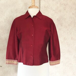 May include: A burgundy button-down shirt with three-quarter sleeves displayed on a mannequin. The cuffs feature a decorative trim with gold, beige, and red patterns. The shirt has a classic collar and is made of a shiny fabric.