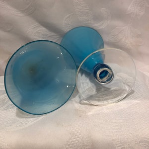 May include: Three blue-tinted glass goblets are arranged on a white patterned surface. The goblets have a clear base and a light blue bowl. The stems are a darker shade of blue.