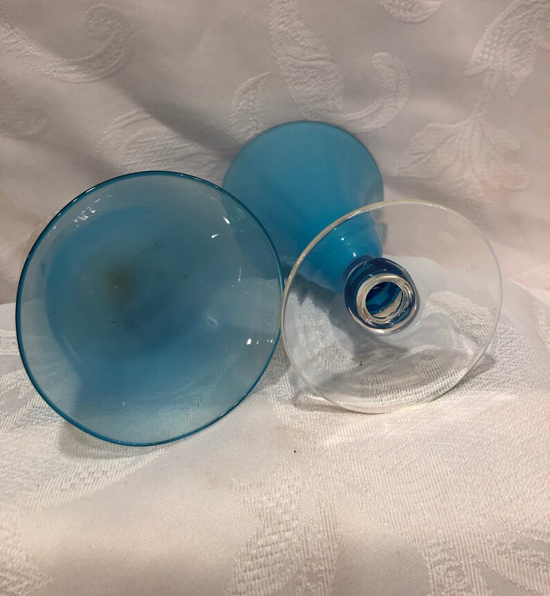 May include: Three blue-tinted glass goblets are arranged on a white patterned surface. The goblets have a clear base and a light blue bowl. The stems are a darker shade of blue.