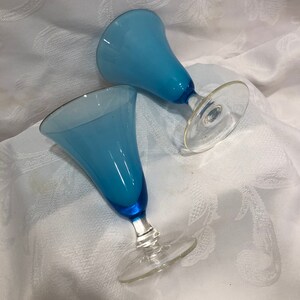 May include: Two blue-tinted glass goblets with clear stems and bases. The goblets have a flared, bell-shaped bowl and a dark blue base. The rims of the goblets have a thin gold trim. The goblets are set on a white, patterned surface.