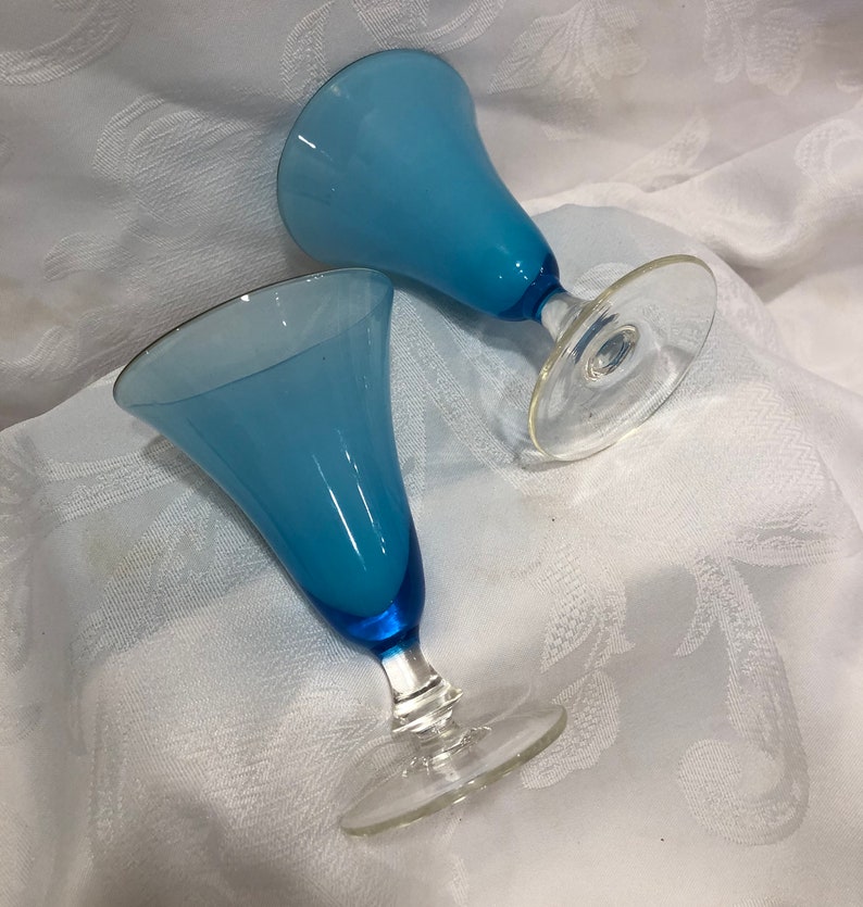 May include: Two blue-tinted glass goblets with clear stems and bases. The goblets have a flared, bell-shaped bowl and a dark blue base. The rims of the goblets have a thin gold trim. The goblets are set on a white, patterned surface.