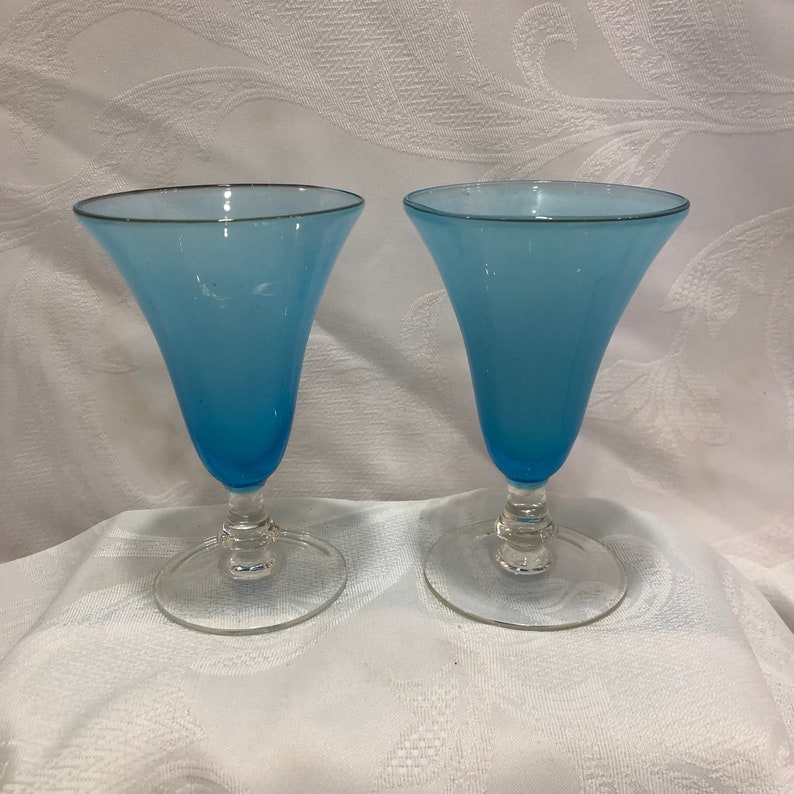 May include: Two vintage blue glass goblets with a flared top and clear stem. The rim of each glass has a silver trim. The goblets are displayed on a white surface.