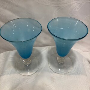 May include: Two blue-tinted glass goblets with a flared top and a clear, round base. The stem has a decorative, twisted design. The goblets are set against a white, textured background.