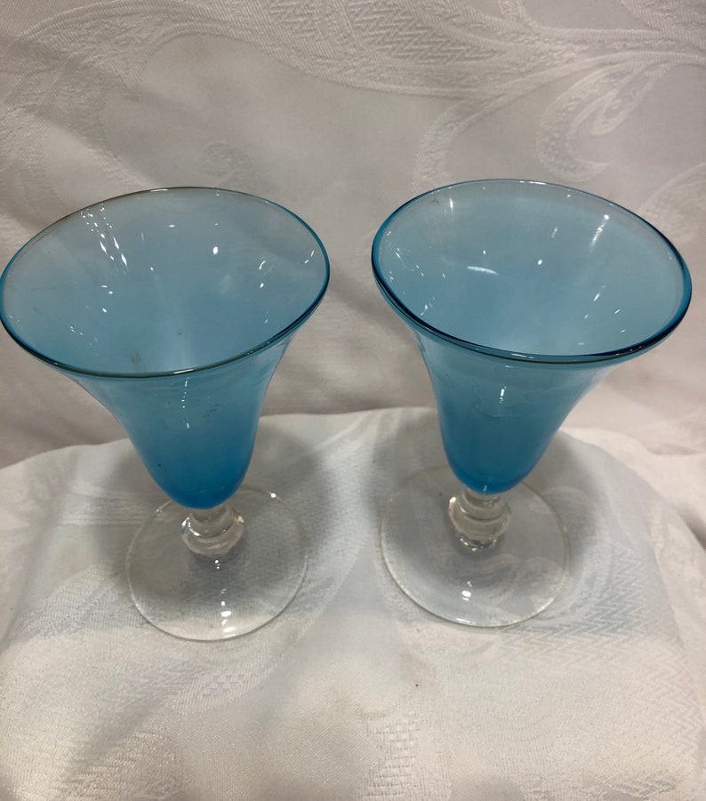 May include: Two blue-tinted glass goblets with a flared top and a clear, round base. The stem has a decorative, twisted design. The goblets are set against a white, textured background.