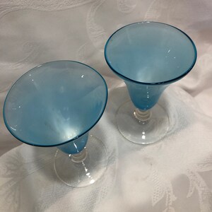 May include: Two light blue stemmed glasses with a flared bowl and clear glass base. The glasses are set against a white background with a subtle floral pattern. The glasses are empty.