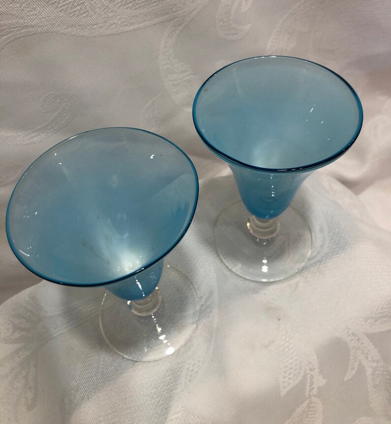 May include: Two light blue stemmed glasses with a flared bowl and clear glass base. The glasses are set against a white background with a subtle floral pattern. The glasses are empty.