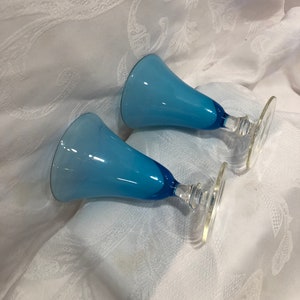 May include: Two blue glass goblets with a flared top and a clear stem and base. The goblets have a gradient of light to dark blue. The stem and base are clear glass.