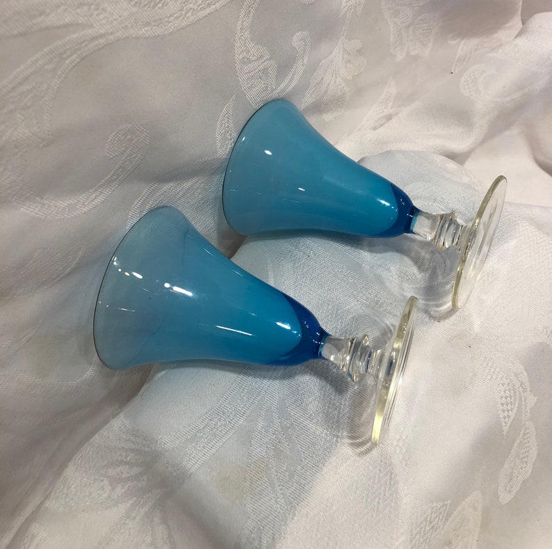 May include: Two blue glass goblets with a flared top and a clear stem and base. The goblets have a gradient of light to dark blue. The stem and base are clear glass.