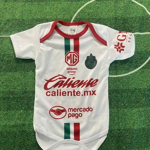May include: White baby bodysuit with red and green accents, featuring the text "Caliente caliente.mx" and "mercado pago". The bodysuit has a round neck and short sleeves. The design includes a team logo and the Amazon Prime logo.