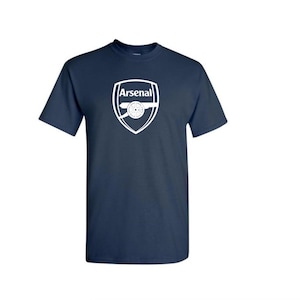 May include: Navy blue t-shirt with the Arsenal logo in white. The logo is a shield shape with the word "Arsenal" at the top and a cannon in the centre. The shirt has short sleeves and a crew neck.