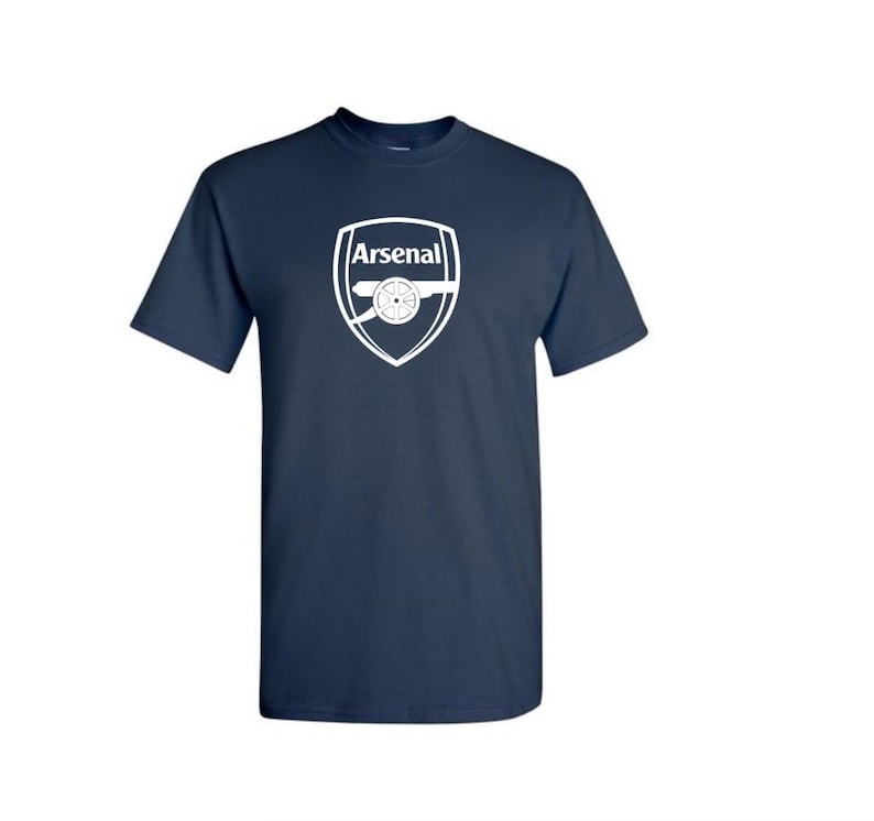 May include: Navy blue t-shirt with the Arsenal logo in white. The logo is a shield shape with the word "Arsenal" at the top and a cannon in the centre. The shirt has short sleeves and a crew neck.