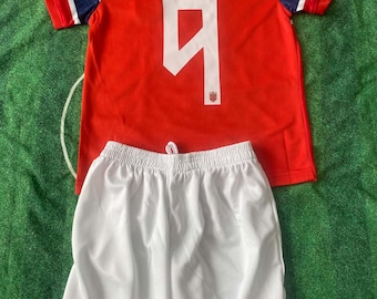 May include: A red soccer jersey with blue and white sleeve accents, featuring the text BRAUT HAALAND and the number 9 in white on the back, paired with plain white athletic shorts with an elastic waistband and a drawstring.
