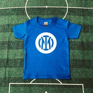 May include: A blue short-sleeved t-shirt with a white circular logo. The logo features the letters "I", "M", and "I" in a stylized design. The shirt is made of a soft material and is suitable for children. The shirt is displayed on a green surface.