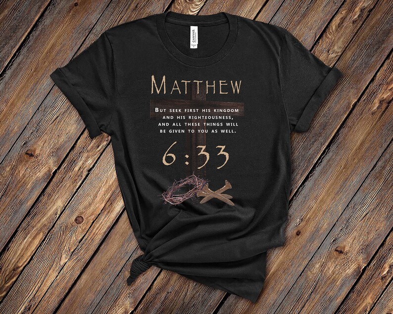 May include: Black t-shirt with a white graphic design featuring the text "MATTHEW" and a bible verse, "BUT SEEK FIRST HIS KINGDOM AND HIS RIGHTEOUSNESS, AND ALL THESE THINGS WILL BE GIVEN TO YOU AS WELL. 6:33". The graphic also includes a stylized cross with nails and a crown of thorns.