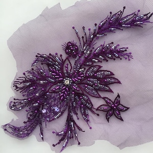 Op de afbeelding: Paarse parelversierde bloemenapplicatie met een strasssteen in het midden. Deze versiering is perfect om kleding, accessoires of woonaccessoires een vleugje glans en elegantie te geven.