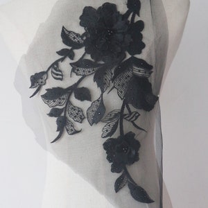 Könnte beinhalten: Schwarze florale Spitzenapplikation mit Perlendetails. Die Applikation zeigt eine große Blume mit kleineren Blumen und Blättern. Die Applikation befindet sich auf einem durchsichtigen Netzstoff.