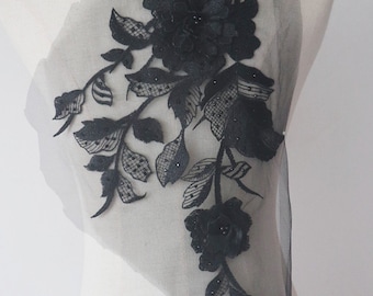 Puede incluir: Aplique de encaje floral negro con detalles de cuentas. El aplique presenta una flor grande con flores y hojas más pequeñas. El aplique está sobre un fondo de malla transparente.