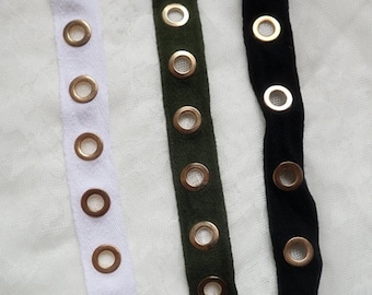 May include: A close-up shot of three fabric strips: white, dark green, and black. Each strip is embellished with regularly spaced, gold-coloured eyelets. These eyelets offer a decorative and practical feature, ideal for diverse craft and design applications.