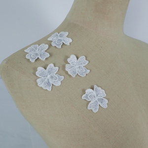 May include: Five white floral lace appliques. Each applique features a detailed flower design with multiple petals. The appliques are made of a delicate, textured fabric and are ideal for embellishing clothing or craft projects.