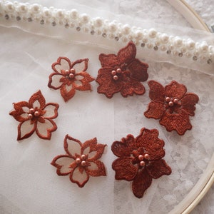 Puede incluir: Seis apliques de flores bordadas de color marrón con centros de perlas. Los apliques están sobre un fondo de encaje blanco.