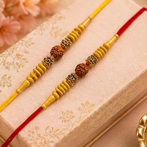 Puede incluir: Dos pulseras decorativas con cuentas rudraksha marrones centrales flanqueadas por espaciadores dorados estriados y pequeños detalles con incrustaciones de cristal. Una banda tiene un hilo amarillo y la otra un hilo rojo, sobre una caja.