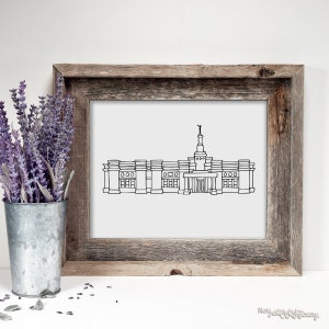 May include: A framed black and white line drawing of a building with a tall spire. The artwork is in a rustic, weathered wooden frame. A galvanized metal bucket with lavender flowers is to the left of the frame.