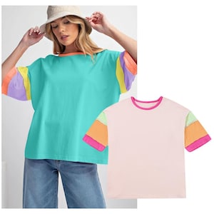 May include: Two short-sleeved t-shirts with colourful striped sleeves. The first shirt is turquoise with purple, yellow, and orange stripes. The second shirt is pink with green, orange, and pink stripes.