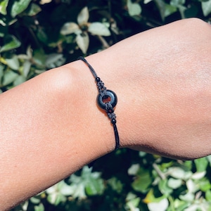 May include: A black bracelet featuring a circular, polished stone bead at the center. The bead is connected to a black cord with adjustable knots. The bracelet is shown against a background of green foliage.