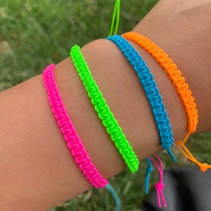 May include: Four neon colored friendship bracelets in pink, green, blue, and orange. The bracelets are tied together and worn on a wrist.