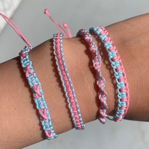May include: Four friendship bracelets in pink and light blue colors. The bracelets are woven together in a simple pattern.