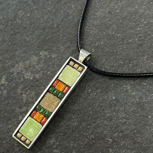 Pode incluir: Um colar com pingente retangular com um design de mosaico. O pingente tem uma moldura prateada e apresenta azulejos verdes, laranja e bege. O colar tem um cordão preto.