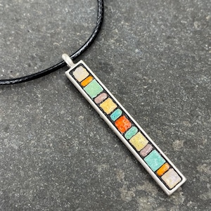 May include: A silver pendant necklace with a rectangular design. The pendant features a mosaic of colorful, square-shaped tiles in shades of orange, green, yellow, and blue. The necklace has a black cord.