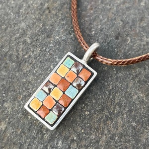 Puede incluir: Colgante rectangular con un diseño de mosaico en tonos naranja, marrón, azul y amarillo, engastado en un marco plateado. El colgante cuelga de un cordón trenzado marrón. El collar se exhibe sobre una superficie gris.