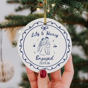 May include: A round white ceramic ornament featuring a dark blue decorative border and a central illustration of a couple. The text reads Lily & Harry, Engaged, and the date 12.13.26. A gold string is attached to the top for hanging purposes.