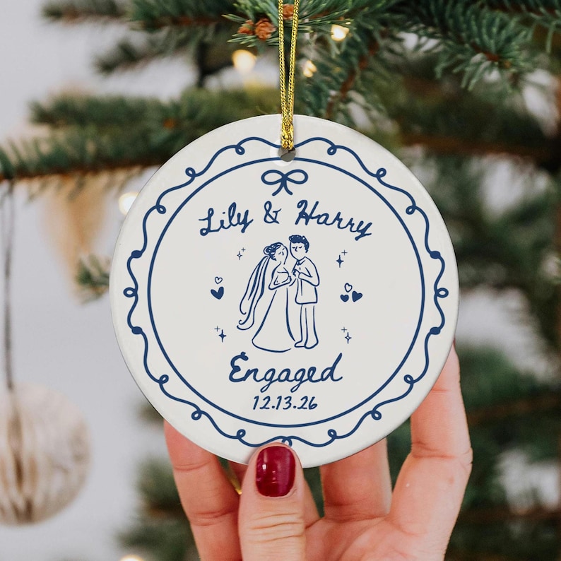 May include: A round white ceramic ornament featuring a dark blue decorative border and a central illustration of a couple. The text reads Lily & Harry, Engaged, and the date 12.13.26. A gold string is attached to the top for hanging purposes.