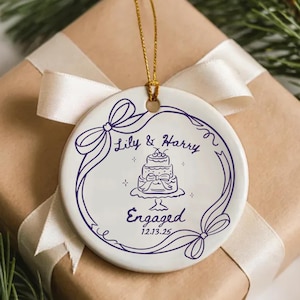 May include: A round white ceramic ornament featuring a dark purple line drawing of a tiered cake, a ribbon border, and the text Lily & Harry Engaged 12.13.25. The ornament hangs from a gold string and is centered on a gift box with a white ribbon.