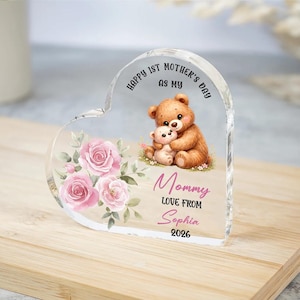 May include: A heart-shaped acrylic keepsake with a teddy bear design, pink roses, and the text "Happy 1st Mother's Day as my Mommy Love From Sophia 2026". The clear acrylic sits on a wooden surface.