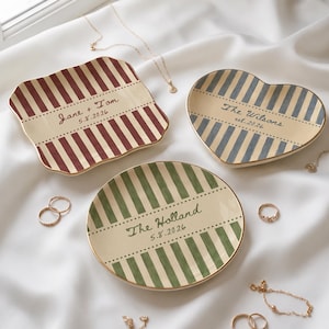 May include: Three ceramic trinket dishes with gold-toned rims. One square dish features red stripes and the text Jane + Tom 5.8.2026. One heart-shaped dish has blue stripes and the text The Wilsons est. 2026. One round dish has green stripes and The Holland 5.8.2026.