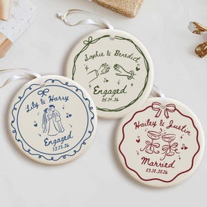 May include: Three round white ceramic ornaments with white ribbon hangers. Each features a decorative border and custom text: one with a couple and 'Lily & Harry Engaged 12.13.26', one with hands and 'Sophie & Benedict Engaged 05.14.26', and one with glasses and 'Hailey & Justin Married 03.10.2026'.