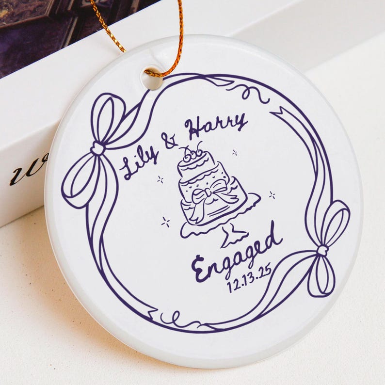 May include: A round white ceramic ornament featuring a dark purple line drawing of a tiered cake, a ribbon border, and the text Lily & Harry, Engaged, and 12.13.25. A gold string is attached through a hole at the top for hanging purposes.