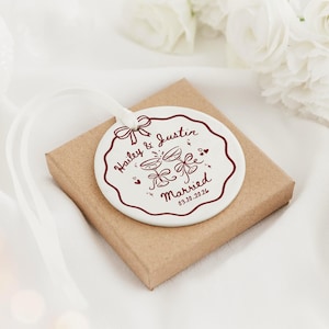May include: A round white ceramic ornament with a scalloped edge, featuring a dark red printed design of two champagne glasses, a bow, and the text Hailey & Justin Married 03.10.2026. A white ribbon is attached to the top of the ornament.