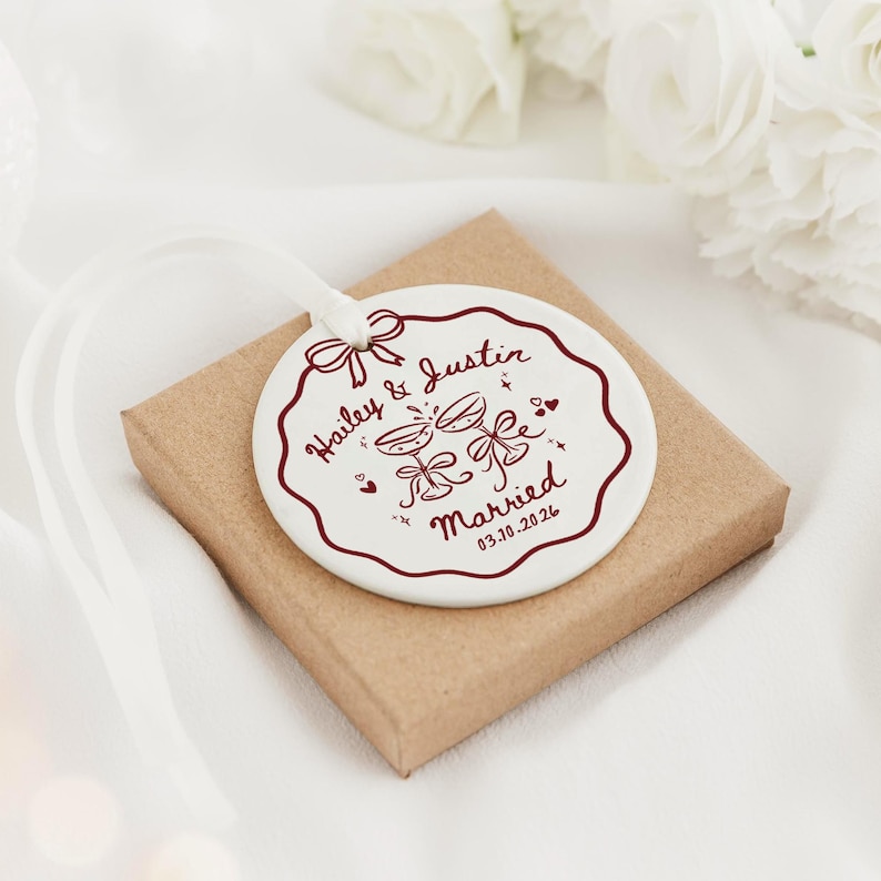 May include: A round white ceramic ornament with a scalloped edge, featuring a dark red printed design of two champagne glasses, a bow, and the text Hailey & Justin Married 03.10.2026. A white ribbon is attached to the top of the ornament.
